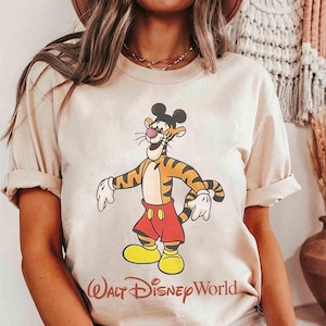 May include: Beige t-shirt featuring a cartoon tiger character with Mickey Mouse ears, red shorts, and yellow shoes. The tiger has orange and black stripes. The text "Walt Disney World" is printed in red script at the bottom.