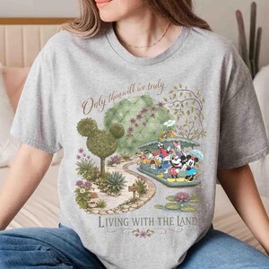 May include: A gray t-shirt featuring a colorful graphic of Disney characters on a boat ride through a garden. The text "Only then will we truly" and "Living with the Land" are also displayed on the shirt.