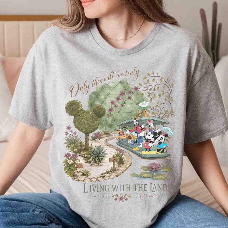 May include: A gray t-shirt featuring a colorful graphic of Disney characters on a boat ride through a garden. The text "Only then will we truly" and "Living with the Land" are also displayed on the shirt.