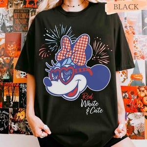 Op de afbeelding: Zwart T-shirt met een cartoonfiguur in rood, wit en blauw, inclusief hartvormige zonnebril en een strik. De tekst "Red White & Cute" is ook zichtbaar.
