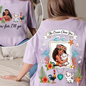 Op de afbeelding: Lichtpaars T-shirt met een cartoon illustratie van een vrouw, een varken en een haan. De voorkant van het shirt heeft de tekst "How far I'll go". De achterkant van het shirt zegt "The Ocean Chose You".