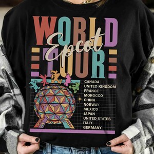 K&ouml;nnte beinhalten: Schwarzes T-Shirt mit einer bunten Grafik des Epcot World Showcase. Die Grafik enth&auml;lt den Text "World Epcot Tour" und eine Liste der im World Showcase vertretenen L&auml;nder: Kanada, Vereinigtes K&ouml;nigreich, Frankreich, Marokko, China, Norwegen, Mexiko, Japan, Vereinigte Staaten, Italien und Deutschland.