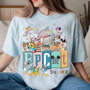 May include: Light blue t-shirt featuring a colorful graphic of Disney characters and landmarks. The word "EPCOT" is displayed in large, colorful letters, with "Since 1982" below. The design includes Mickey Mouse, Minnie Mouse, Donald Duck, and Goofy.
