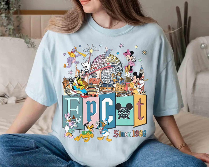 May include: Light blue t-shirt featuring a colorful graphic of Disney characters and landmarks. The word "EPCOT" is displayed in large, colorful letters, with "Since 1982" below. The design includes Mickey Mouse, Minnie Mouse, Donald Duck, and Goofy.