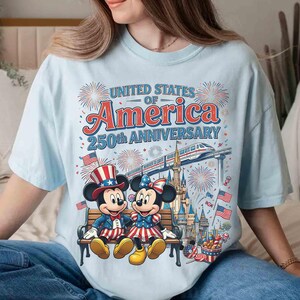 Może przedstawiać: Jasnoniebieska koszulka z grafiką Myszki Miki i Myszki Minnie w patriotycznych strojach, upamiętniająca 250. rocznicę Stan&oacute;w Zjednoczonych Ameryki. Projekt zawiera fajerwerki, flagi i pociąg, z napisem "United States of America 250th Anniversary."