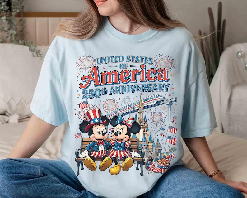 Może przedstawiać: Jasnoniebieska koszulka z grafiką Myszki Miki i Myszki Minnie w patriotycznych strojach, upamiętniająca 250. rocznicę Stan&oacute;w Zjednoczonych Ameryki. Projekt zawiera fajerwerki, flagi i pociąg, z napisem "United States of America 250th Anniversary."