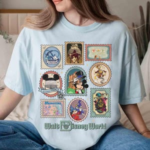 Puede incluir: Camiseta azul claro con un collage de coloridos dise&ntilde;os de sellos postales con varios personajes y logotipos de Disney. El texto "Walt Disney World" est&aacute; impreso en la parte inferior de la camiseta.