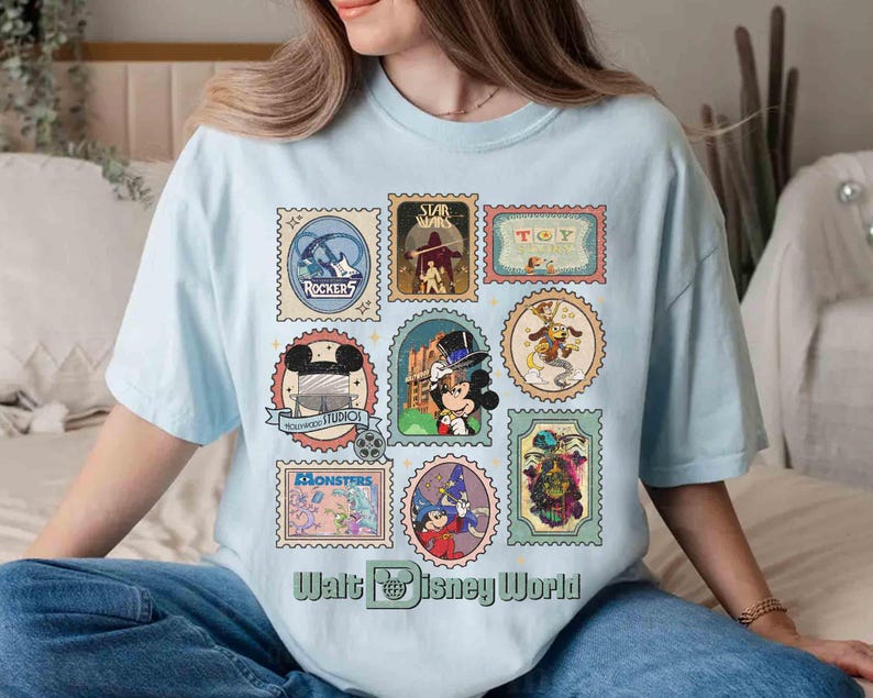 Puede incluir: Camiseta azul claro con un collage de coloridos dise&ntilde;os de sellos postales con varios personajes y logotipos de Disney. El texto "Walt Disney World" est&aacute; impreso en la parte inferior de la camiseta.