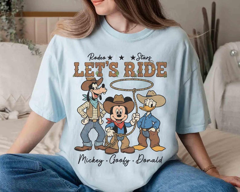 May include: Light blue t-shirt featuring a graphic of Mickey Mouse, Goofy, and Donald Duck dressed as cowboys with the text "Rodeo Stars LET'S RIDE" and the names "Mickey, Goofy, Donald" below the characters.