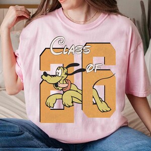 May include: A light pink t-shirt featuring a cartoon image of Pluto the dog inside a large orange number 26. The words "CLASS OF" are arched above the number. The shirt is a crew neck style.