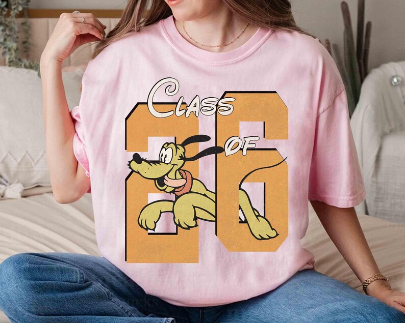 May include: A light pink t-shirt featuring a cartoon image of Pluto the dog inside a large orange number 26. The words "CLASS OF" are arched above the number. The shirt is a crew neck style.