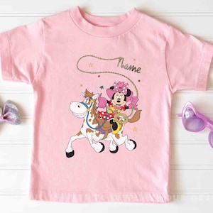 May include: A light pink t-shirt featuring a cartoon Minnie Mouse riding a horse with a lasso. Minnie Mouse is wearing a pink cowgirl hat and outfit. The word "Name" is written above the image in a cursive font. The shirt has a round neck.