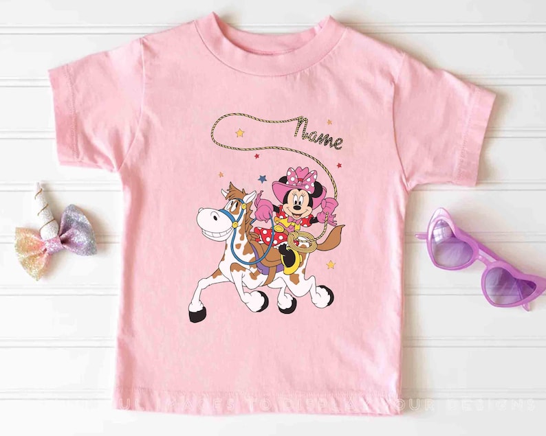 May include: A light pink t-shirt featuring a cartoon Minnie Mouse riding a horse with a lasso. Minnie Mouse is wearing a pink cowgirl hat and outfit. The word "Name" is written above the image in a cursive font. The shirt has a round neck.