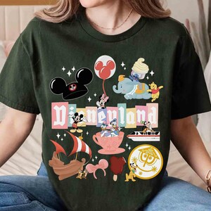 May include: Dark green t-shirt featuring a colorful Disneyland graphic. The design includes Mickey Mouse ears, Minnie Mouse, Dumbo, Winnie the Pooh, and other iconic Disney characters. The word "Disneyland" is prominently displayed.