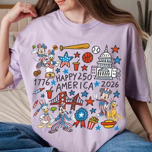 Puede incluir: Una camiseta de color lila claro con un gr&aacute;fico colorido de personajes de dibujos animados y s&iacute;mbolos patri&oacute;ticos. El dise&ntilde;o incluye a Mickey y Minnie Mouse, el Pato Donald y Pluto, con el texto "Happy 250 America 2026" y el a&ntilde;o "1776".