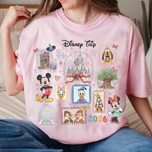 Puede incluir: Camiseta rosa con un dise&ntilde;o de Disney Trip. El dise&ntilde;o incluye varios personajes de Disney como Mickey Mouse, Minnie Mouse, Donald Duck y Pluto, junto con el texto "2026". La camiseta tiene cuello redondo y mangas cortas.