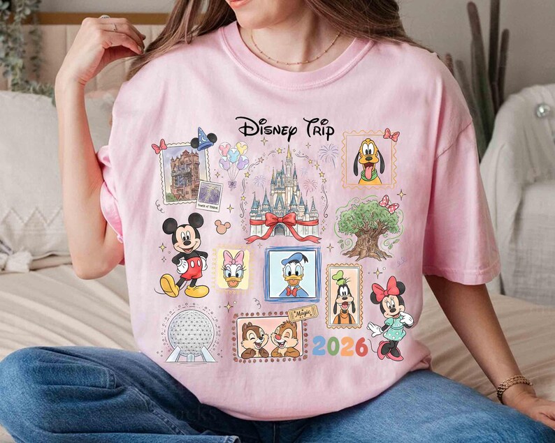 Puede incluir: Camiseta rosa con un dise&ntilde;o de Disney Trip. El dise&ntilde;o incluye varios personajes de Disney como Mickey Mouse, Minnie Mouse, Donald Duck y Pluto, junto con el texto "2026". La camiseta tiene cuello redondo y mangas cortas.