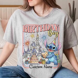 May include: A gray t-shirt featuring a colorful graphic with the text "BIRTHDAY Boy" and "Custom Name". The graphic includes a cartoon character, a castle, fireworks, a cake, and wrapped gifts.