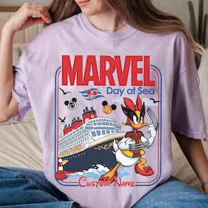 May include: A light purple t-shirt featuring a graphic design with the words "MARVEL Day at Sea". The design includes a cruise ship, cartoon characters, and the text "Custom Name".