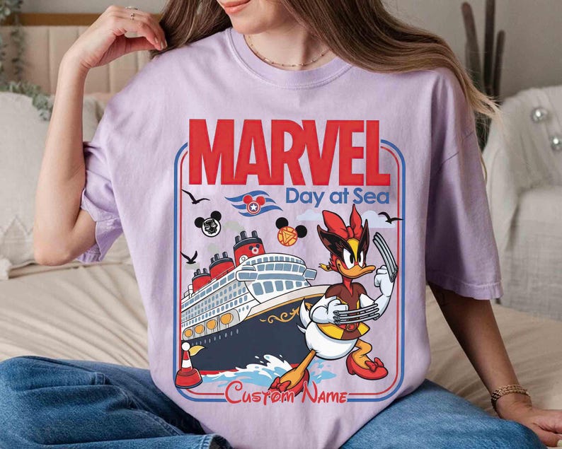 May include: A light purple t-shirt featuring a graphic design with the words "MARVEL Day at Sea". The design includes a cruise ship, cartoon characters, and the text "Custom Name".