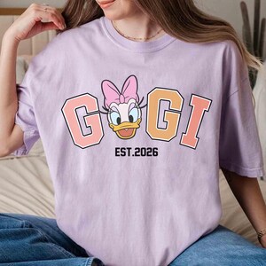 May include: A light purple t-shirt featuring a cartoon image of Daisy Duck with the text "GIGI" in a collegiate style font. Below the text is "EST.2026" in black. The shirt is a casual style.