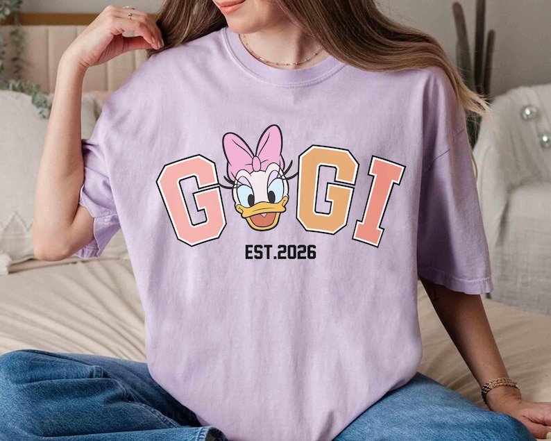 May include: A light purple t-shirt featuring a cartoon image of Daisy Duck with the text "GIGI" in a collegiate style font. Below the text is "EST.2026" in black. The shirt is a casual style.