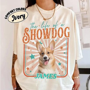 May include: Ivory-colored t-shirt with the text "the life of a SHOWDOG" in orange and teal. A smiling corgi dog is centered in a rectangular frame with the name "JAMES" below. The shirt also has the words "COMFORT COLORS" and "Ivory" in an oval.