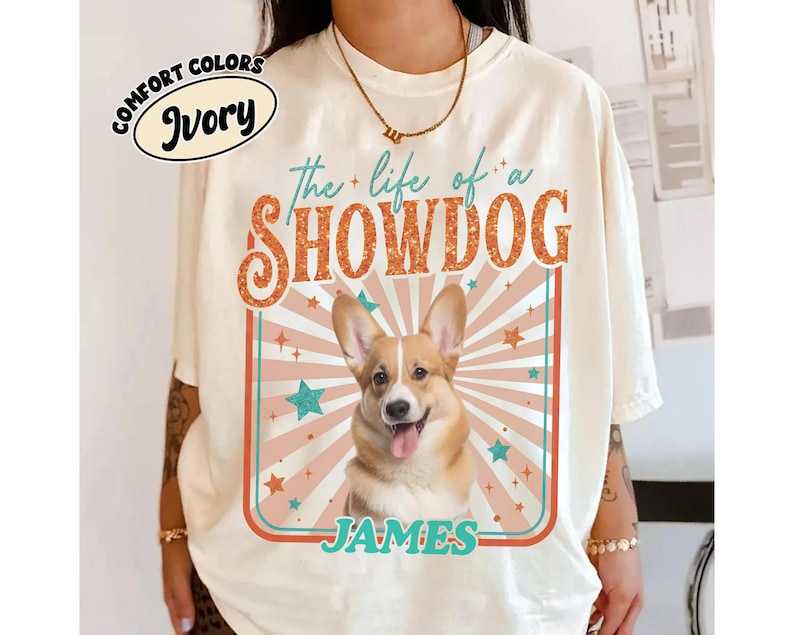May include: Ivory-colored t-shirt with the text "the life of a SHOWDOG" in orange and teal. A smiling corgi dog is centered in a rectangular frame with the name "JAMES" below. The shirt also has the words "COMFORT COLORS" and "Ivory" in an oval.