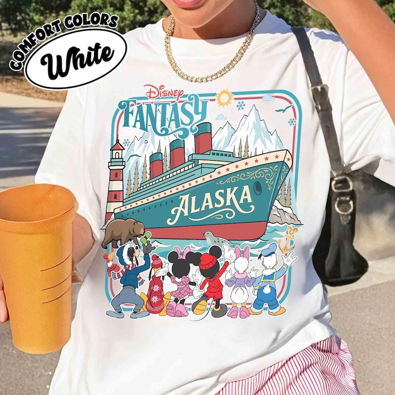 May include: White t-shirt with a Disney Fantasy Alaska graphic. The design features a cruise ship, mountains, and cartoon characters. The shirt has the words "Comfort Colors" and "White" in a circular design.