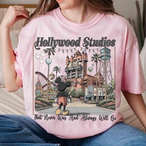 K&ouml;nnte beinhalten: Hellrosa T-Shirt mit einem Grafikdesign von Hollywood Studios und Mickey Mouse. Das Design enth&auml;lt den Text "Hollywood Studios" und "That Never Was And Always Will Be". Das Bild zeigt eine Themenpark-Szene mit einer Achterbahn und einem Wasserturm.