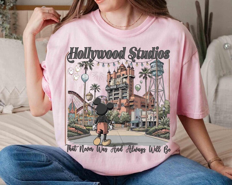 K&ouml;nnte beinhalten: Hellrosa T-Shirt mit einem Grafikdesign von Hollywood Studios und Mickey Mouse. Das Design enth&auml;lt den Text "Hollywood Studios" und "That Never Was And Always Will Be". Das Bild zeigt eine Themenpark-Szene mit einer Achterbahn und einem Wasserturm.