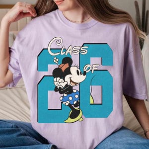 May include: A light purple t-shirt featuring a graphic of Minnie Mouse and the text "CLASS OF 26" in a teal blue color. Minnie Mouse is wearing a polka dot skirt and yellow shoes.