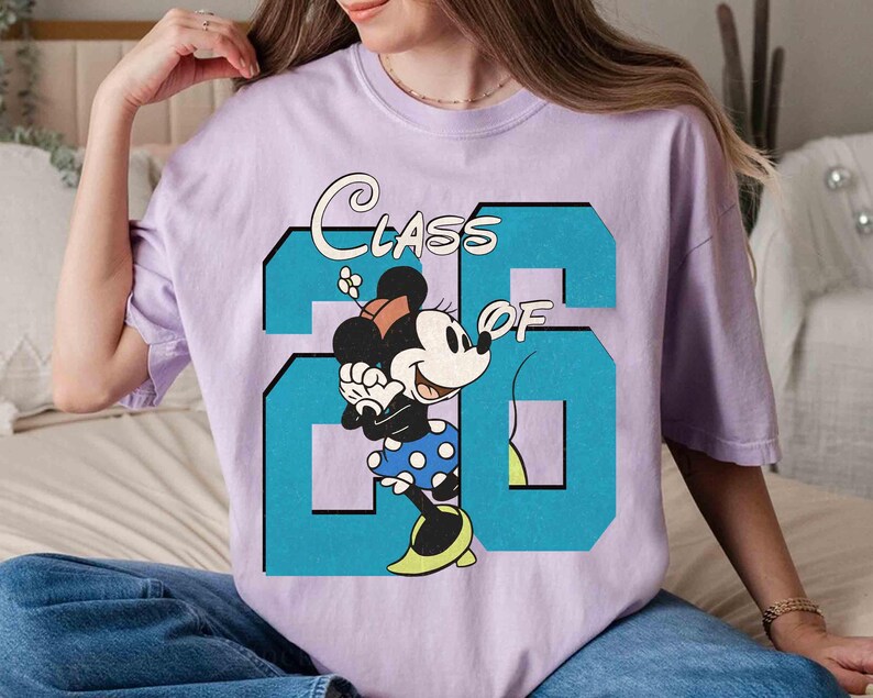 May include: A light purple t-shirt featuring a graphic of Minnie Mouse and the text "CLASS OF 26" in a teal blue color. Minnie Mouse is wearing a polka dot skirt and yellow shoes.