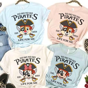 May include: Four t-shirts with a pirate theme. The shirts are light blue, pink, white, and blue. Each shirt features a cartoon character wearing a pirate hat and holding a sword. The text on the shirts reads "Yo Ho Yo Ho Pirates Est. 2024 Life For Me".