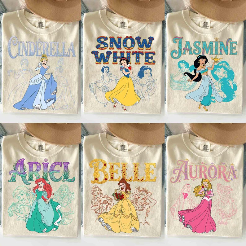 Op de afbeelding: Zes cr&egrave;mekleurige t-shirts met Disney-prinsessen. Elk shirt heeft de naam van een prinses in een kleurrijk lettertype, samen met een illustratie van de prinses. De prinsessen zijn Assepoester, Sneeuwwitje, Jasmine, Ariel, Belle en Aurora.
