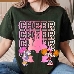 May include: Dark green t-shirt featuring the word "CHEER" stacked three times with a pink castle silhouette. Daisy Duck and Minnie Mouse cheerleaders are on either side of the castle, holding pom-poms. The shirt is a comfortable, casual style.