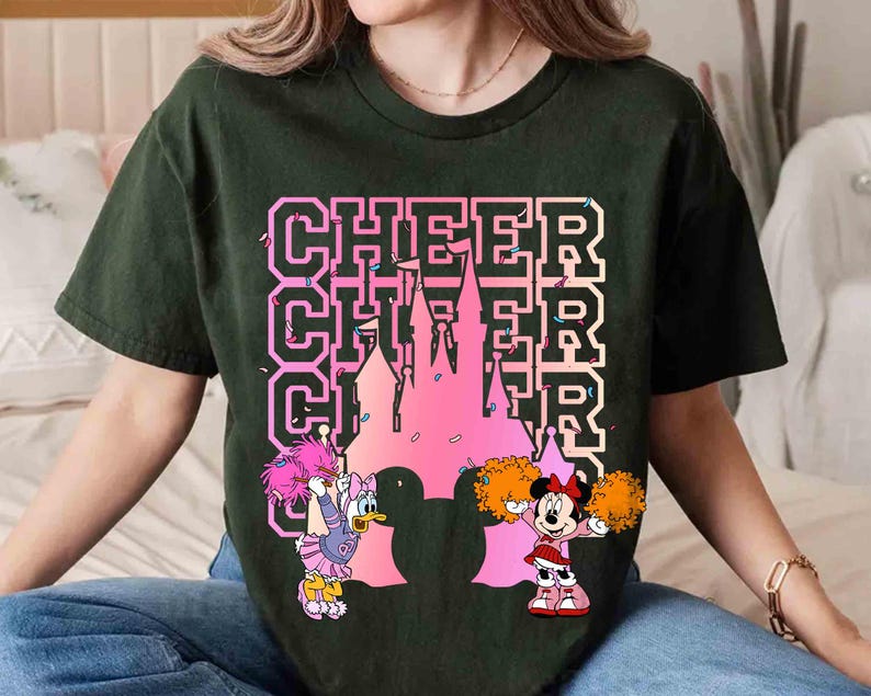 May include: Dark green t-shirt featuring the word "CHEER" stacked three times with a pink castle silhouette. Daisy Duck and Minnie Mouse cheerleaders are on either side of the castle, holding pom-poms. The shirt is a comfortable, casual style.