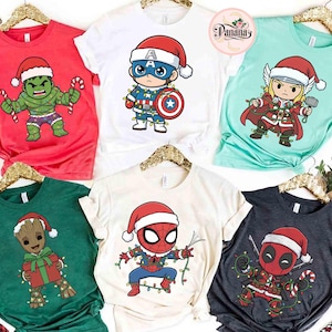 May include: Collection of Christmas-themed t-shirts featuring Marvel superheroes. Designs include Hulk, Captain America, Thor, Groot, Spider-Man, and Deadpool, all wearing Santa hats and festive lights. Shirts are in various colors, including red, white, green, and charcoal.