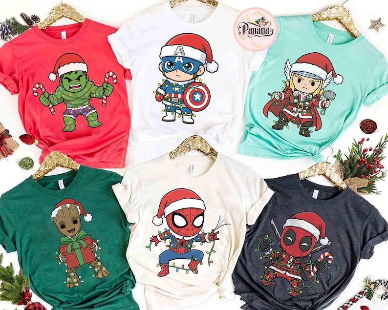 May include: Collection of Christmas-themed t-shirts featuring Marvel superheroes. Designs include Hulk, Captain America, Thor, Groot, Spider-Man, and Deadpool, all wearing Santa hats and festive lights. Shirts are in various colors, including red, white, green, and charcoal.