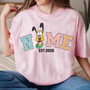 May include: A light pink t-shirt featuring the word "NOME" in a collegiate font, with a cartoon dog character. The shirt includes the text "EST.2026" below the word "NOME".