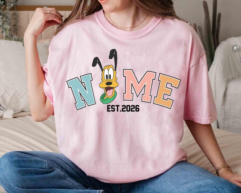 May include: A light pink t-shirt featuring the word "NOME" in a collegiate font, with a cartoon dog character. The shirt includes the text "EST.2026" below the word "NOME".
