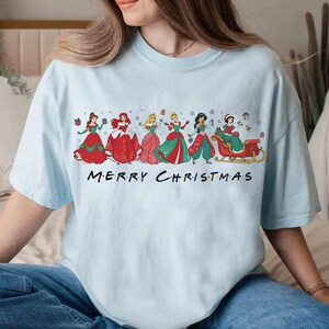 May include: Light blue t-shirt featuring a festive design with several cartoon princesses in holiday attire. The phrase "MERRY CHRISTMAS" is printed in black block letters across the bottom of the graphic. The shirt is made of a soft material.