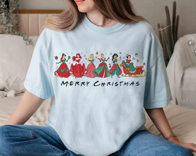 May include: Light blue t-shirt featuring a festive design with several cartoon princesses in holiday attire. The phrase "MERRY CHRISTMAS" is printed in black block letters across the bottom of the graphic. The shirt is made of a soft material.