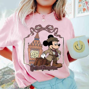 May include: Pink t-shirt featuring a graphic of Mickey Mouse in a travel outfit, standing in front of Big Ben. The words "United Kingdom" are below the image. A white mug with a smiley face is held in the hand.