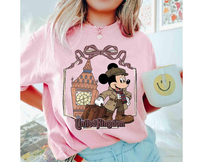 May include: Pink t-shirt featuring a graphic of Mickey Mouse in a travel outfit, standing in front of Big Ben. The words "United Kingdom" are below the image. A white mug with a smiley face is held in the hand.