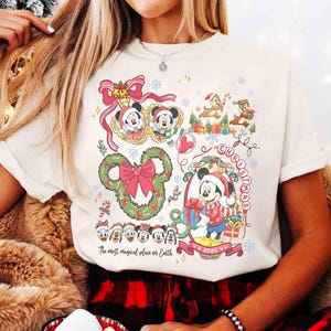 May include: Cream-colored t-shirt featuring a festive Christmas design with Mickey Mouse and friends. The graphic includes a Mickey Mouse-shaped wreath, the words "Christmas" and "2025", and the phrase "The most magical place on Earth".