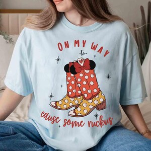 Peut inclure: T-shirt bleu clair avec un motif de bottes &agrave; pois rouges et blancs avec un n&oelig;ud rouge. Le texte sur le t-shirt est "On my way to cause some ruckus".