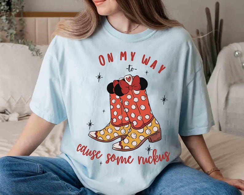 Peut inclure: T-shirt bleu clair avec un motif de bottes &agrave; pois rouges et blancs avec un n&oelig;ud rouge. Le texte sur le t-shirt est "On my way to cause some ruckus".