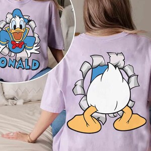 May include: A light purple t-shirt featuring a cartoon graphic of Donald Duck bursting through a torn paper effect. The front of the shirt displays Donald with the name "DONALD". The back shows Donald's rear.