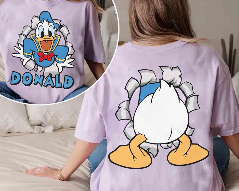 May include: A light purple t-shirt featuring a cartoon graphic of Donald Duck bursting through a torn paper effect. The front of the shirt displays Donald with the name "DONALD". The back shows Donald's rear.